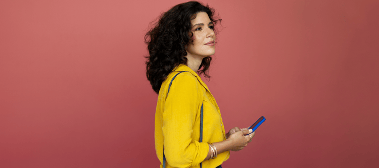 Mulher sorridente com cabelo cacheado, vestindo camisa amarela. Ela segura um celular, contra um fundo rosa. Estilo moderno e vibrante.