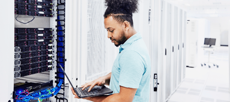 Profissional de TI trabalhando em um datacenter, usando um laptop para monitorar servidores e rede, com cabos coloridos visíveis ao fundo.