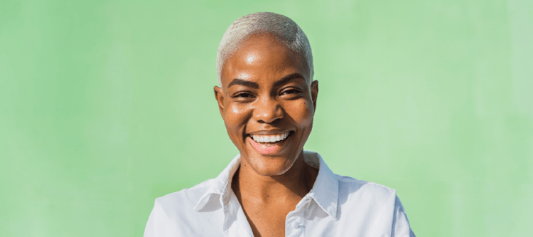 Mulher sorridente com cabelo curto e platinado usando uma camisa branca, em frente a um fundo verde claro, transmitindo alegria e confiança.