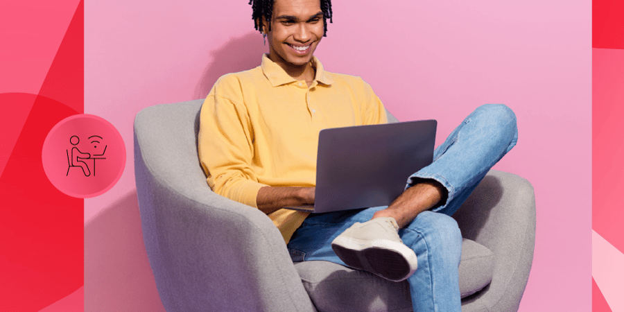 Um jovem sorridente sentado em uma poltrona confortável, usando um laptop, com um fundo rosa vibrante. Ideal para trabalho remoto e inspiração de home office.